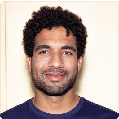 Homem sorridente com cabelo escuro encaracolado e barba veste uma camiseta azul-marinho contra um fundo claro