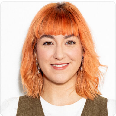 A woman with vibrant orange hair styled with bangs, smiling at the camera. She's wearing a white top and olive green vest