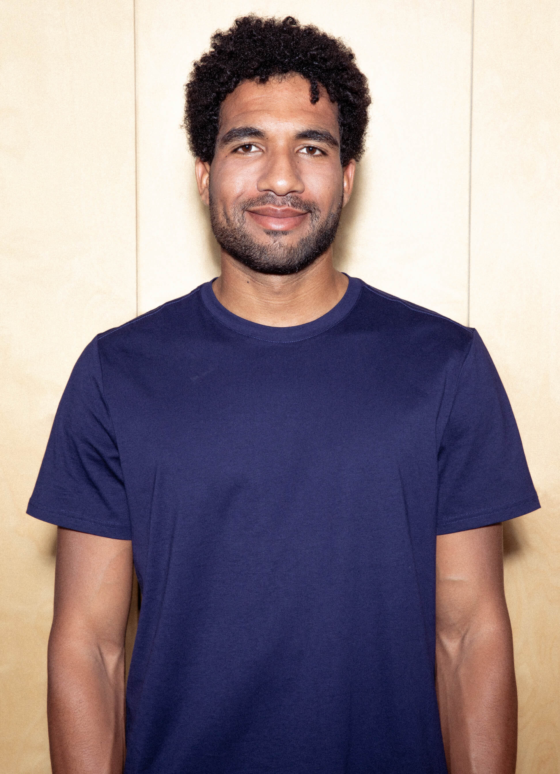 Retrato de homem sorridente com cabelo escuro encaracolado e barba, vestindo uma camiseta azul-marinho contra um fundo colorido claro